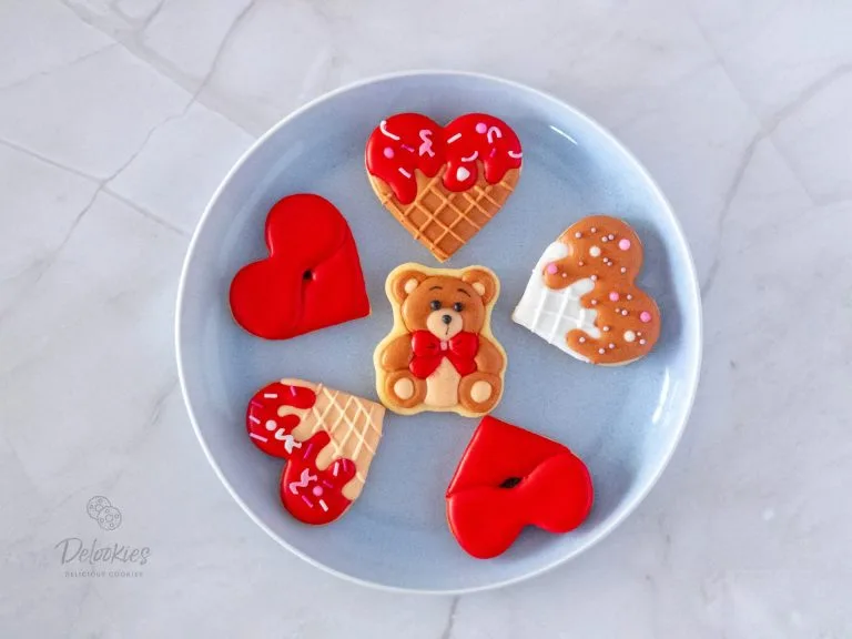 A plate of beautifully crafted Valentine's-themed cookies from Delookies, featuring a charming teddy bear cookie, textured cone-inspired heart cookies with drizzled icing, red lips cookies, and a heart-shaped cookie with sprinkles, all decorated with smooth and decorative icing.