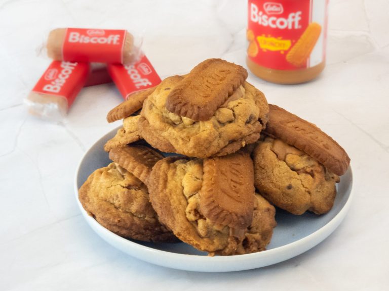 A plate of Deluxe Biscoff Loaded Cookies with Biscoff cream and biscuit packs in the background, showcasing the rich caramelised flavour.