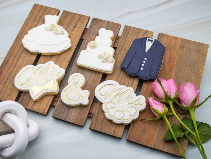 Wedding cookies with Mr, Mrs, wedding gown, and tuxedo cookies flat on a wooden surface surrounded by decorative props.